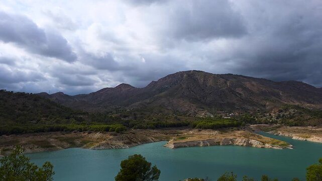 Timelapse footage of dark dramatic storm clouds moving fast over the mountain landscape and turquoise water of Amadorio reservoir in Spain. Ideal for weather, nature, and background videos.