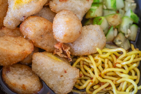 close up of fried fish cake pempek palembang topped with yellow noodles, diced cucumbers, and dark sweet sour sauce.