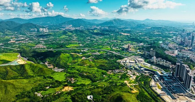 High angle panoramic view shows lush green mountains and modern cityscape in Shenzhen under a clear blue sky with fluffy clouds.