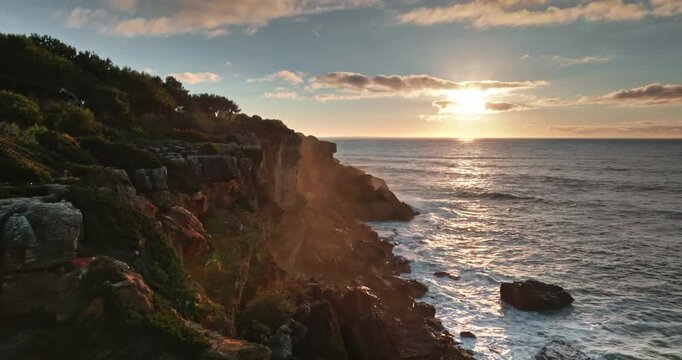 Scenic coastal Cascais landscape at sunset with golden light illuminating rugged cliffs, green vegetation, Atlantic Ocean waves crashing against the shoreline. Nature beauty landscape. Drone flight