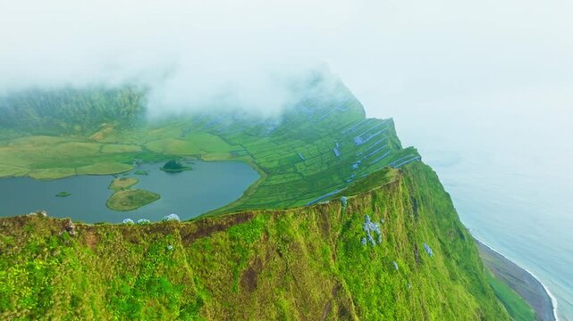 Steep ridge, cliffs, blooming blue hydrangeas and green Caldeirao crater with lakes on Corvo Island, Azores, Portugal. Fog and Clouds. Moving Forward