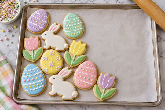 Easter sugar cookies shaped like eggs, rabbits and tulips decorated with pastel royal icing on a baking sheet beside a bowl of sprinkles