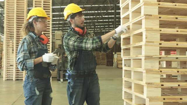 Male and female carpenter team wearing hard hats and safety glasses measure red painted pallet lumber tape measure the organized stacking handling these materials industrial facility classification.