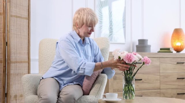 Pretty senior woman sniffing beautiful carnation flowers in armchair at home