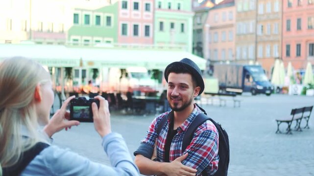 A smiling man and a woman with a camera and backpacks are enjoying a daytime visit in a vibrant historical market square with outdoor cafes and pastel buildings.