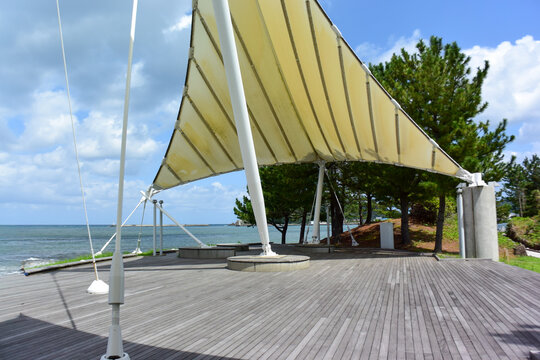 海を見渡す公園に設置された巨大な白いタープテントとウッドデッキ。青空のもとで潮風を感じながら過ごせる開放的な屋外イベントスペースと海辺の景観。兵庫県香住の海岸沿い公園の風景。