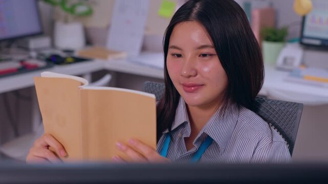 Young Asian adult woman reading book in modern office workplace, seated employee at desk during work break, focused calm expression, computer monitor foreground, gentle smile, quiet concentration