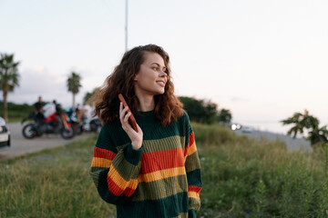 Obraz premium Smiling young woman with curly hair in colorful striped sweater holds smartphone outdoors at sunset. Concept of communication and freedom.