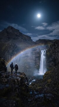 A tiny couple standing beside a quiet waterfall in Iceland under a bright full moon, delicate moonbow forming through the mist, dark volcanic hills surrounding the scene, enormous night sky
