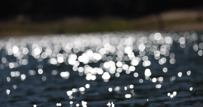 Sunlight glistening with mesmerising abstract sparkles across the surface of a freshwater lake against a dark tree-lined distant shore. Captured in slow motion on a bright sunny day at water level.