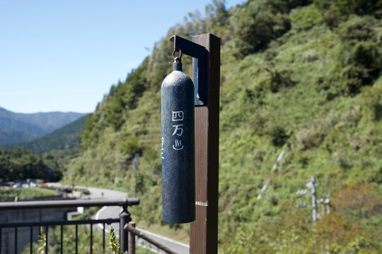 青空と山々を背景に立つ奥四万湖展望台のクマ除けの鈴 Bear warning bell at Okushima Lake viewpoint against blue sky and green mountains