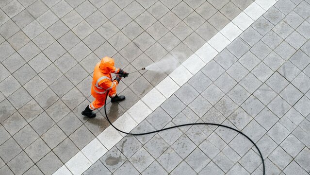 High angle view janitor using pressure washer on tiled floor to clean the street outside office building.