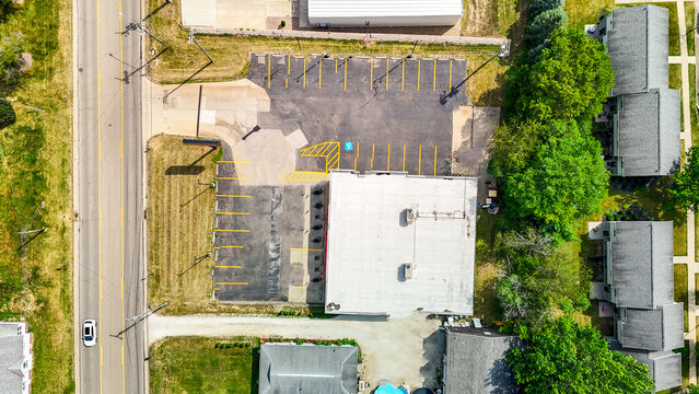 Aerial view of a commercial building with asphalt parking lot and adjacent road. High angle drone perspective of property layout with marked parking spaces. Real estate and urban development