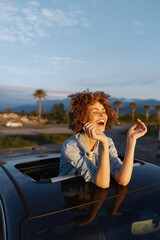 Obraz premium Young woman with curly hair leans out of a car window, smiling brightly on a sunny day, palm trees in the background, denim shirt, carefree road trip mood.