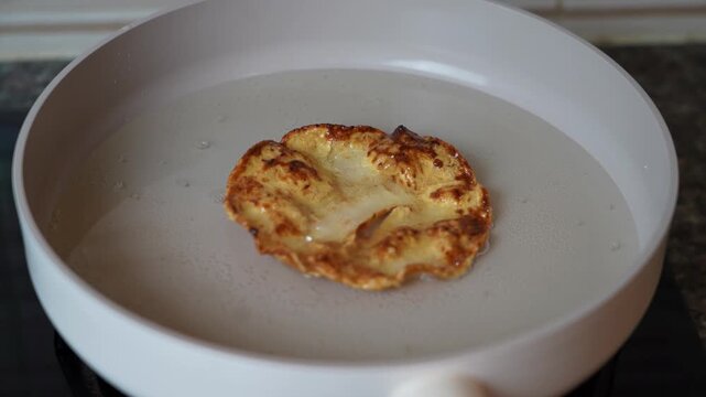 Close up footage of a melinjo cracker frying in a pan during preparation of emping manis, a traditional Indonesian sweet snack. A spatula lifts and turns the cracker in hot oil.
