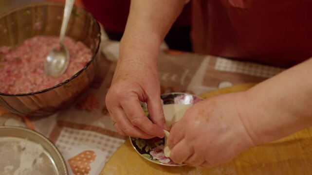 Close up shot captures senior cook skillfully pleating thin dough wrapper over meat filling on floral saucer while making khinkali dumplings in Tbilisi home.