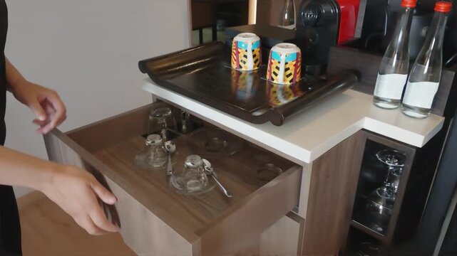 Woman Opening Wooden Drawer with Glassware at Hotel Room Coffee Station.