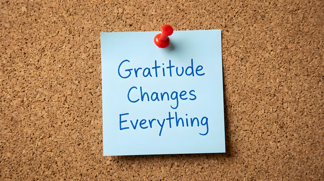 Light blue square sticky note pinned to a textured brown corkboard with a red pushpin featuring handwritten blue ink text reading gratitude changes everything centered in the photographic frame