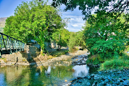 The confluence of the River Colwyn and River Glaslyn with a footbridge framed by lush green trees in Beddgelert village, Snowdonia National Park, North Wales.