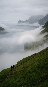 Two tiny silhouettes standing on an empty green hillside in the Faroe Islands, dense ocean fog rolling across the valley and a faint white fogbow forming inside it, distant cliffs barely visible