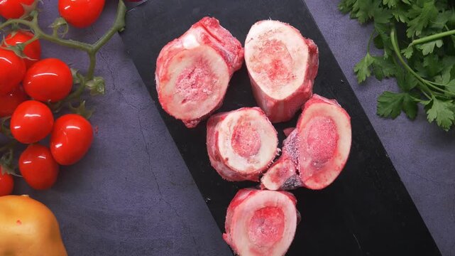 Raw beef marrow bones on a black slate, fresh red tomatoes, and green parsley, ready for cooking bone broth, dynamic food preparation clip.