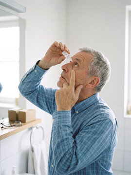 Senior man carefully applying moisturizing eye drops in bathroom mirror