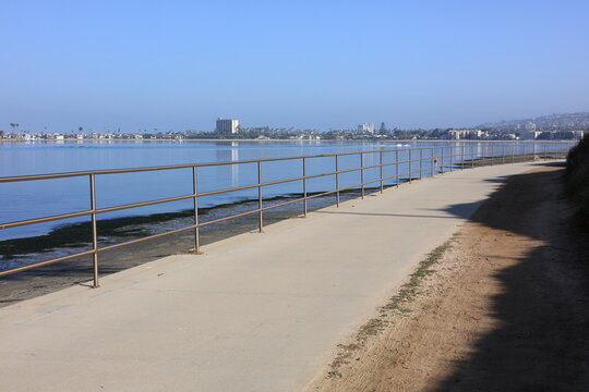 A smooth concrete pedestrian path features a protective metal handrail along the calm bayfront water, with distant buildings under a clear blue sky, San Diego, California