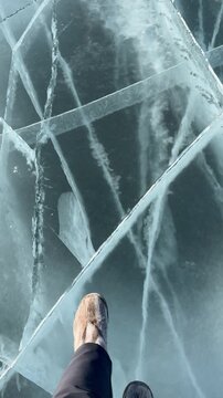 Vertical video of ice texture of frozen Lake Baikal on cold winter day. Female tourist wearing warm fur boots Unty walks across clear, cracked ice. Top view; only her legs are visible. Extreme travels