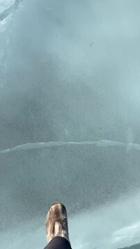 Vertical video of natural ice texture of frozen Lake Baikal on cold winter day. Female tourist wearing warm fur boots Unty walks across clear, cracked ice. Only her legs are visible. Extreme travel