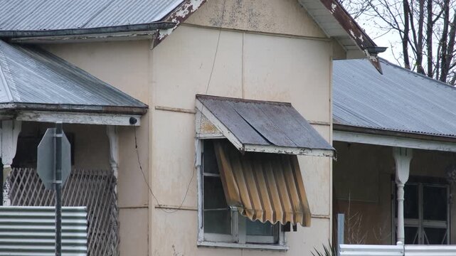 a poorly maintained old house with rusted iron roofs, peeling paint, a corroded and sagging window awning and a worn timber verandah on an rainy day in Australia. Ageing and deteriorating housing