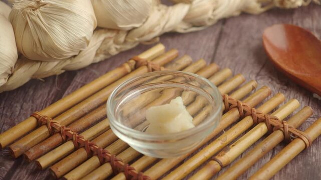 Stop motion animation of crushed garlic paste filling glass bowl on bamboo mat with garlic bulbs background.