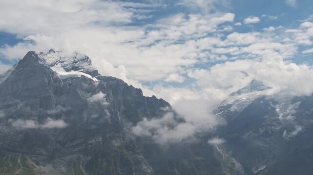 Panoramic view of snow covered Alpine peaks surrounded by clouds in Grindelwald, Switzerland. Mountain landscape with blue sky, sunlight and dramatic clouds. Scenic nature and travel background.