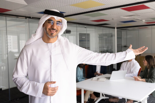 Middle Eastern businessman in kandura stands in conference room, making welcoming gesture to client, inviting partners, colleagues seated at table engaging in professional business office discussion