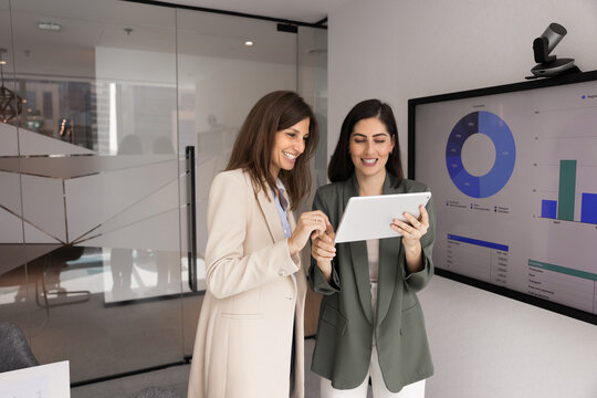 Two smiling multiethnic businesswomen colleagues collaborating on digital tablet in corporate boardroom with visual data charts analyzing statistics discussing strategic insights using fintech tools