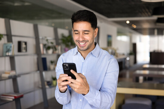 Cheerful young businessman using smartphone, making personal or business call, responding to email, reading message or checking calendar, schedule or social media during workday break in modern office