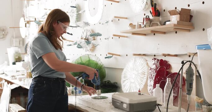 Craftswoman wearing protective glasses using electric hand drilling tool to make hole in glass workpiece performing technical manufacturing task for creating glasswork installation in workshop studio