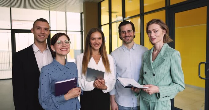 Group portrait diverse coworkers standing together in corporate office looking at camera smiling laughing expressing collaborative team spirit unity professional confidence positive teamwork culture