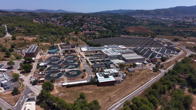 Large wastewater treatment plant with circular clarifier tanks surrounded by industrial buildings, solar panels, and green hills near a suburban town under clear blue sky