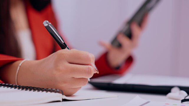 Woman Writing in Notebook with Pen While Holding Tablet at Office Desk, Focused Study and Education Session, Business Planning, Productivity, Digital Communication, Pink Background and Red Sleeve