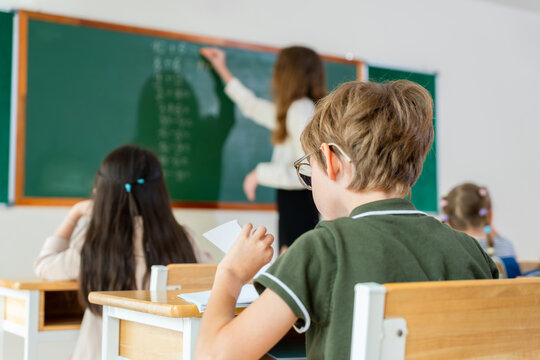 Back view of Students in classroom during a lesson, Primary school teacher writing on chalkboard with diverse pupils in class, Teacher conducting a math lesson in a diverse elementary classroom