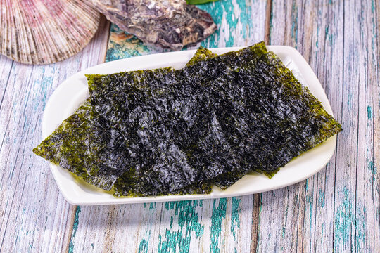 Crispy dark green nori sheets stacked on white rectangular plate with seashell and dried fish on weathered blue and white wooden table, food ingredient