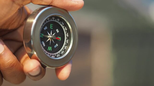 Person holding metal magnetic compass against blurred natural backdrop showing orientation.