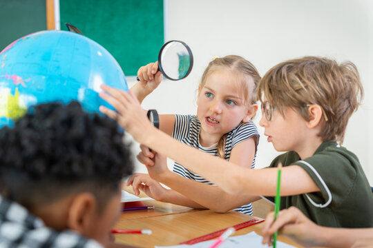Multiethnic school children using magnifying glass to examine globe in classroom, Diverse group of elementary students studying geography, Curious kids learning science and geography in classroom
