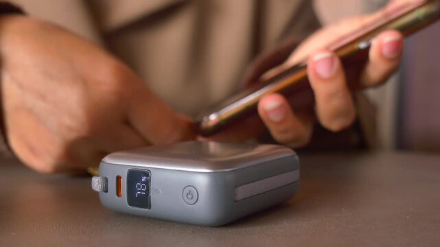 Person uses smartphone near power bank with digital display showing charge level.