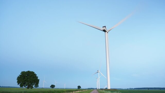 Foreground wind turbine with blurred blades rotating beside gravel road through green farmland under clear blue sky, with wind, turbine, blade, tower detail
