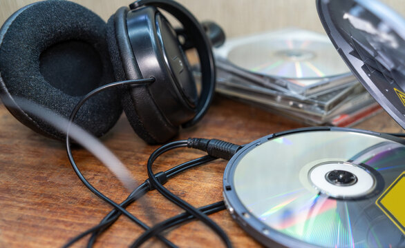 Wired headphones next to CDs and an open floppy drive on a wooden table. Listening to music and playing audio in the style of the 90s. Collection of physical media and digital technologies.
