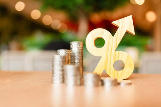 Interest rate increase and inflation rise concept, wooden percentage sign with upward arrow standing beside coin stacks on table, symbolizing monetary tightening, loan growth, and profit.