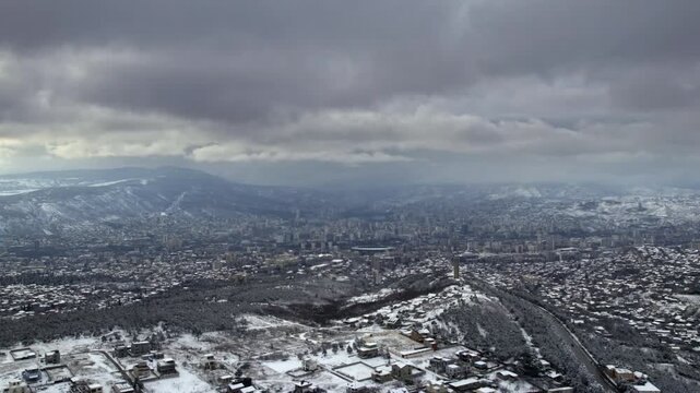 aerial glide above the Tbilisi valley with low houses a solitary tower distant apartment blocks and white mountain slopes snow lined highway bends beside a wooded ridge during a gentle lateral