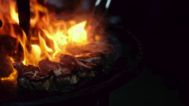 Meat steaks grilling over open fire flames on barbecue grill at night. Cook using tongs to turn roasted beef. Outdoor cooking, fire and smoke, barbecue party concept.