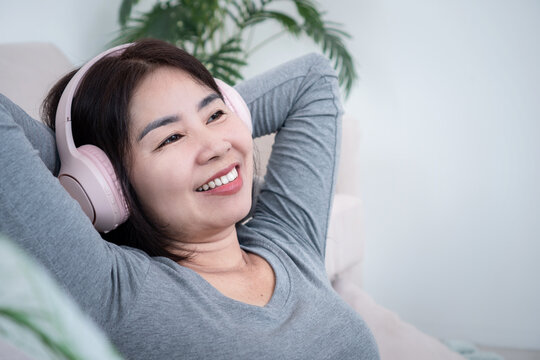 Happy Asian woman relaxing on couch wearing wireless headphones listening to music, smiling, resting head on hands, enjoying leisure audio entertainment, wellness and peaceful home 
